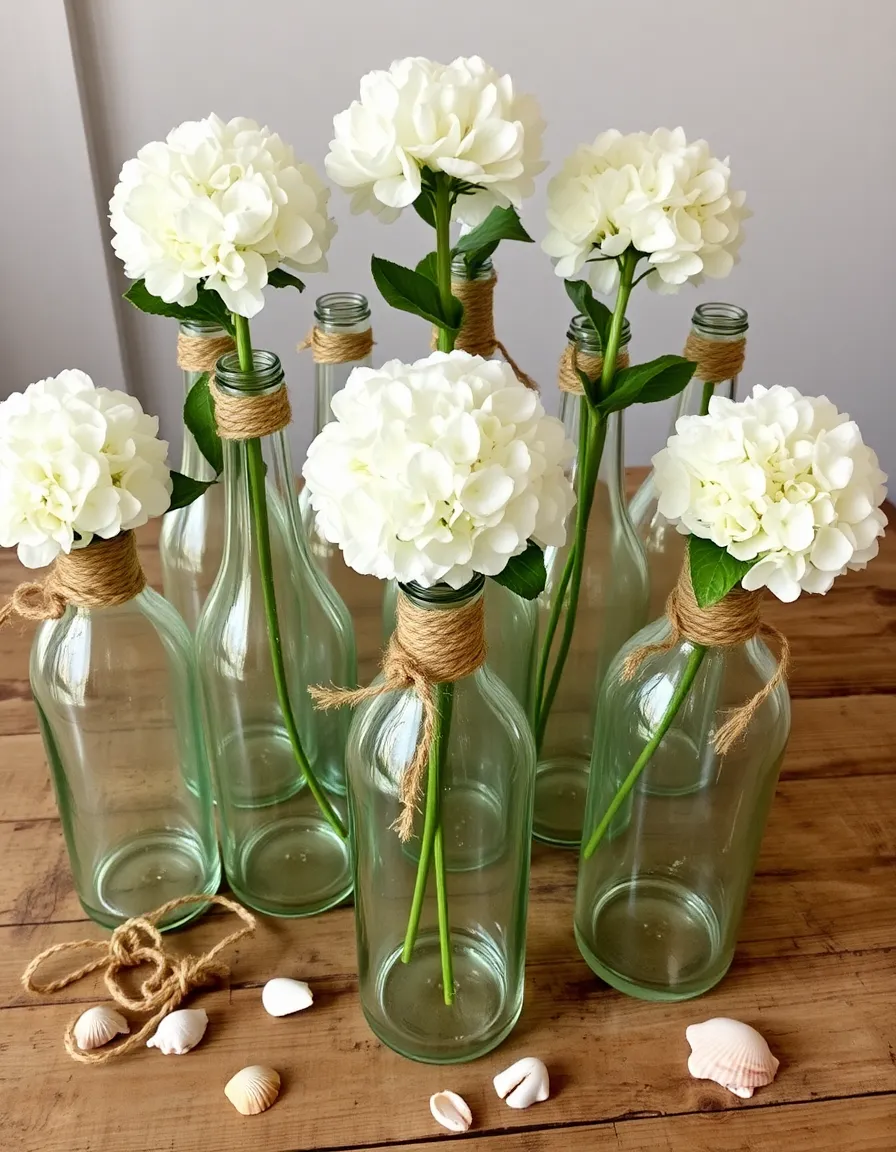 A collection of clear glass bottles with rope-wrapped necks, each holding a single white hydrangea bloom. The bottles are grouped on a weathered wooden table, with a few strands of twine and a handful of seashells scattered around them.