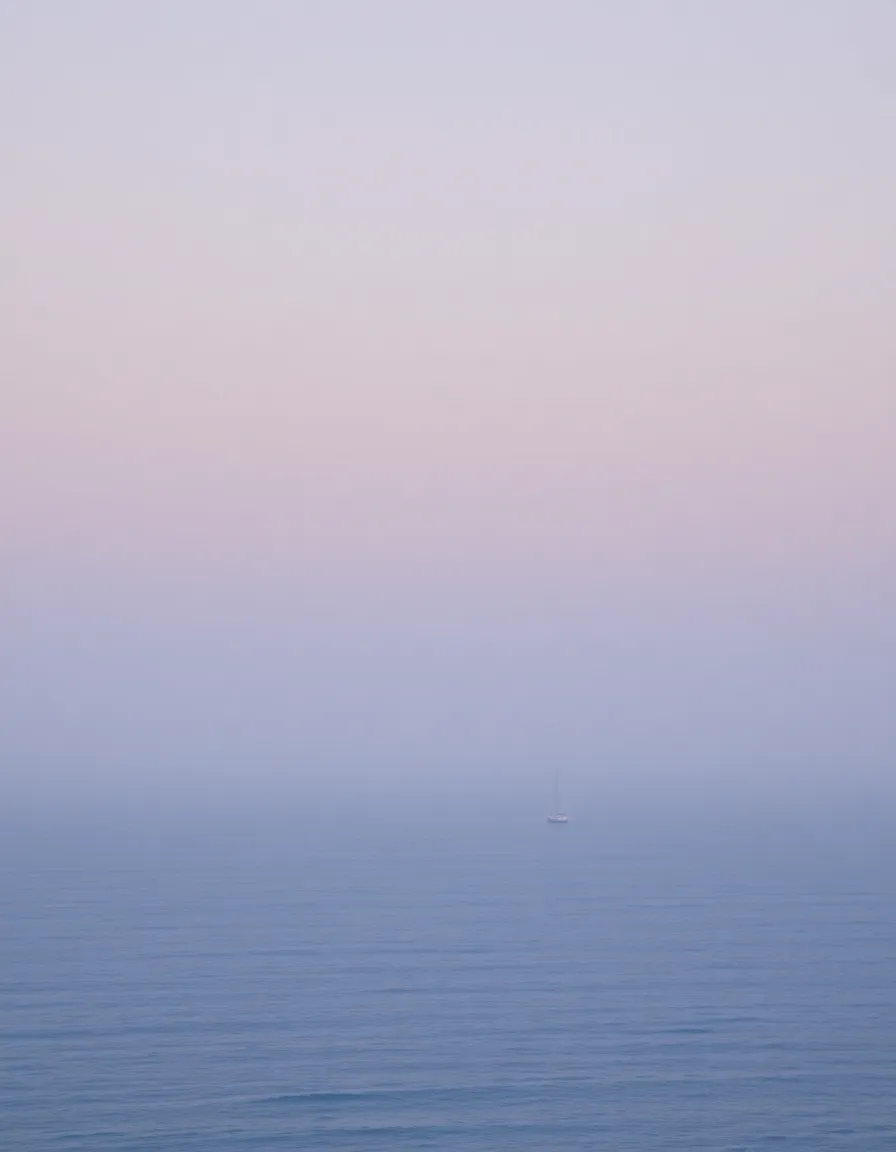 A tranquil seascape at dawn, with a thick mist hovering over calm, silvery-blue water. The horizon blends seamlessly into a pale lavender sky, and a lone sailboat drifts in the distance.